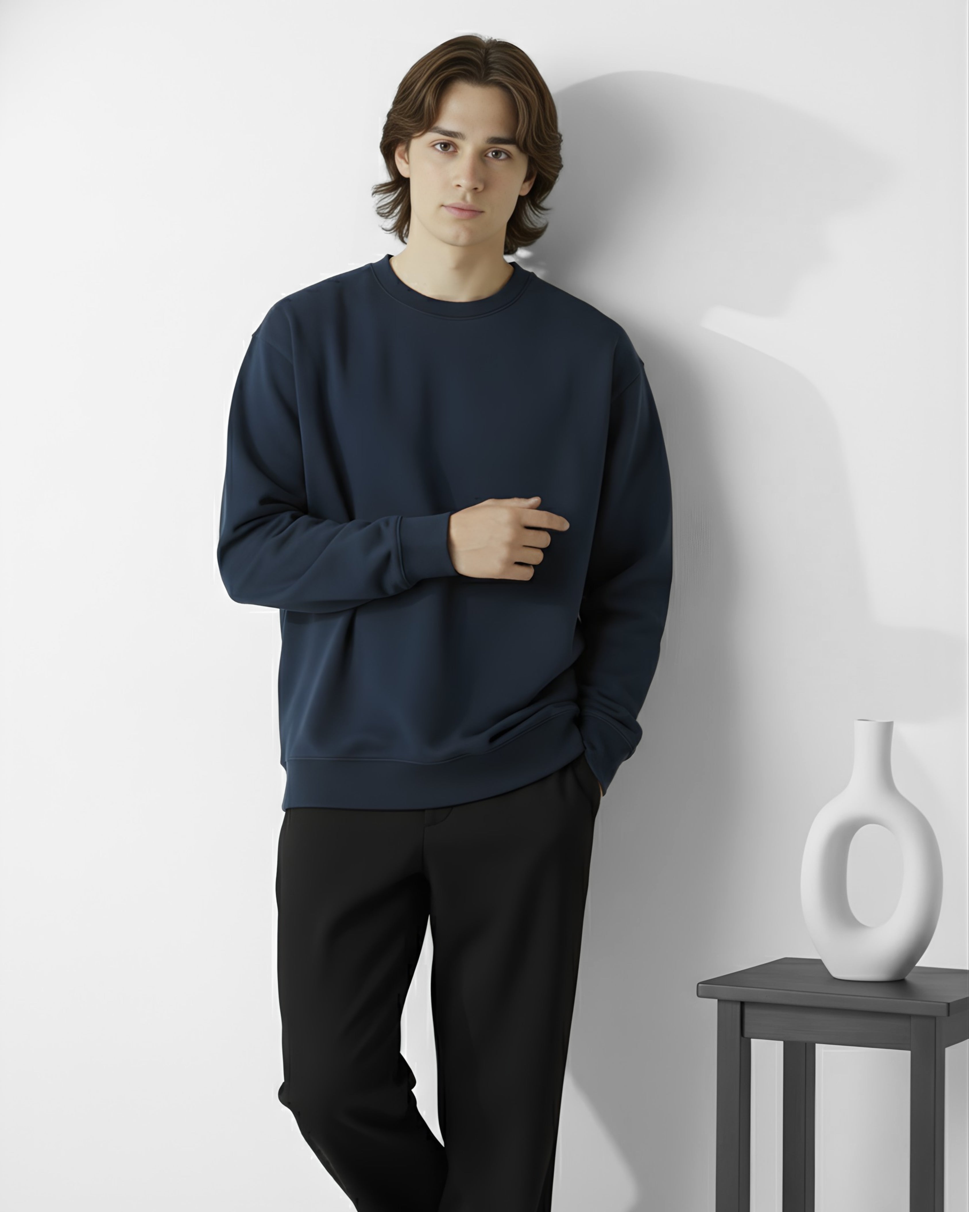Man wearing a navy blue sweatshirt and black pants standing against a white wall with a vase on a table.